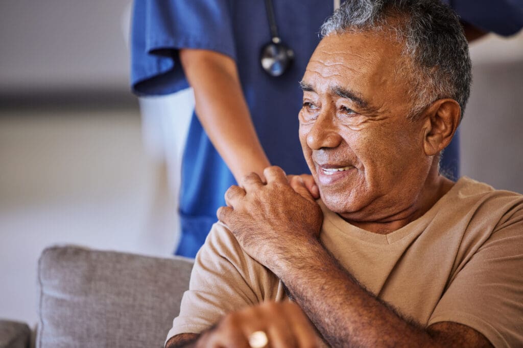 An elderly man clasps the hand of a healthcare worker placed reassuringly on his shoulder.